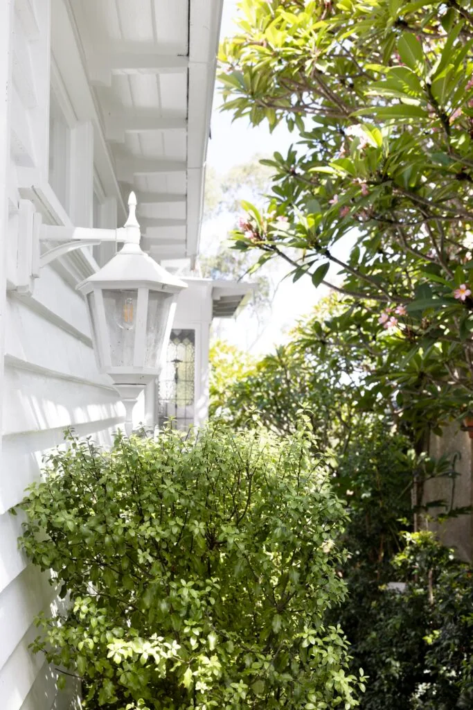 A coach lamp on the side of the home above green bushes.