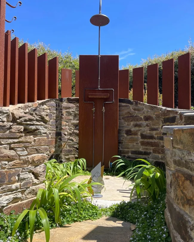 Outdoor shower with stone walls, rust-colored metal panels, and greenery under a clear blue sky.