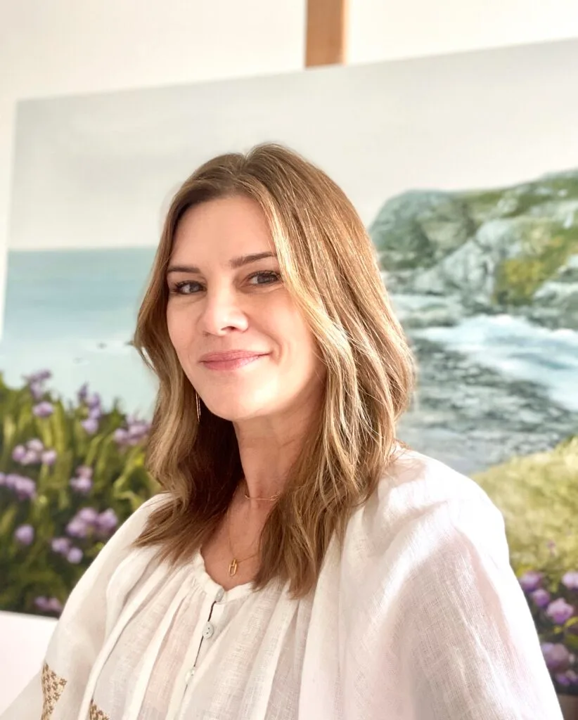 A portrait of artist Annie Hawkins by a coastal landscape painting.