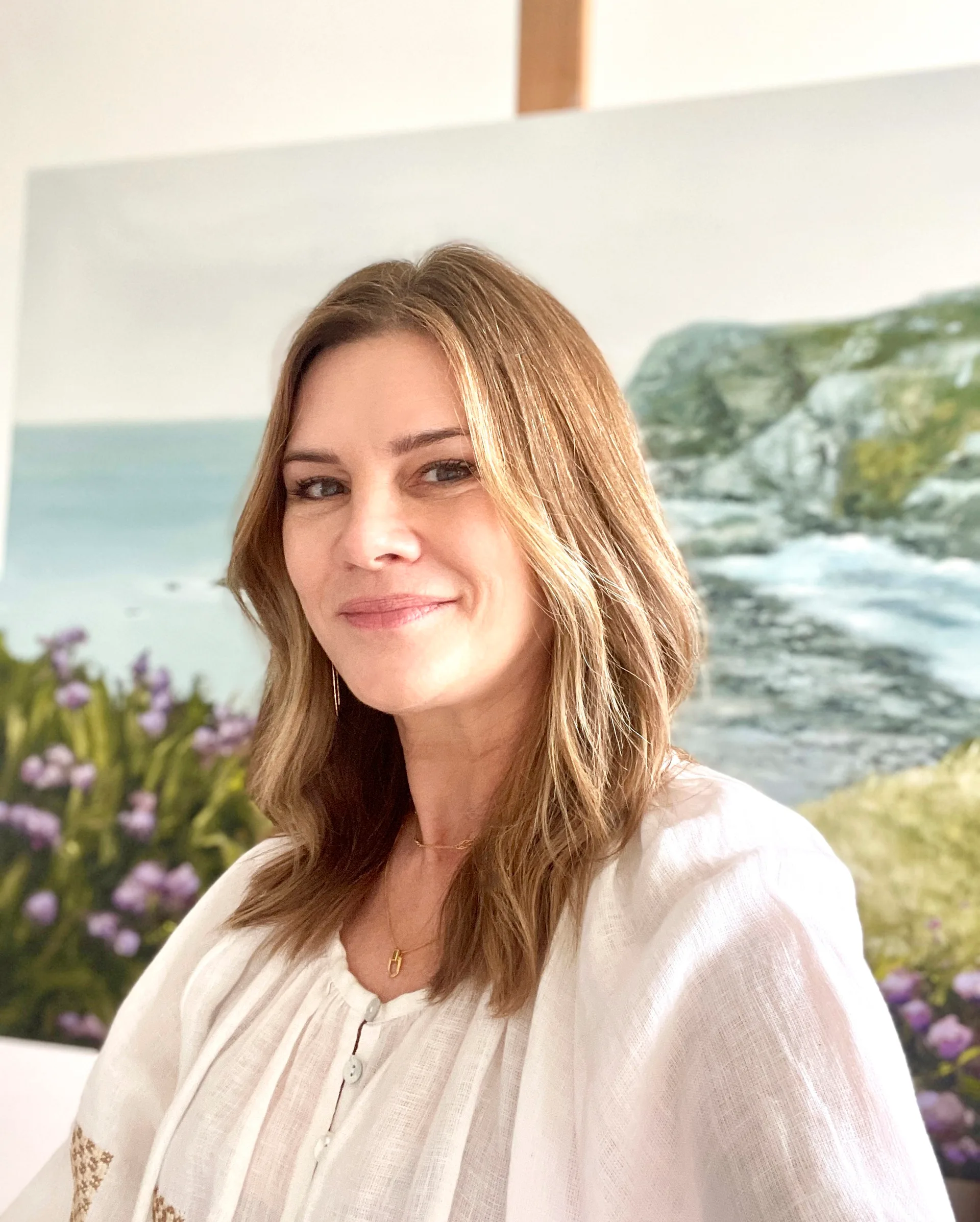 A portrait of artist Annie Hawkins by a coastal landscape painting.