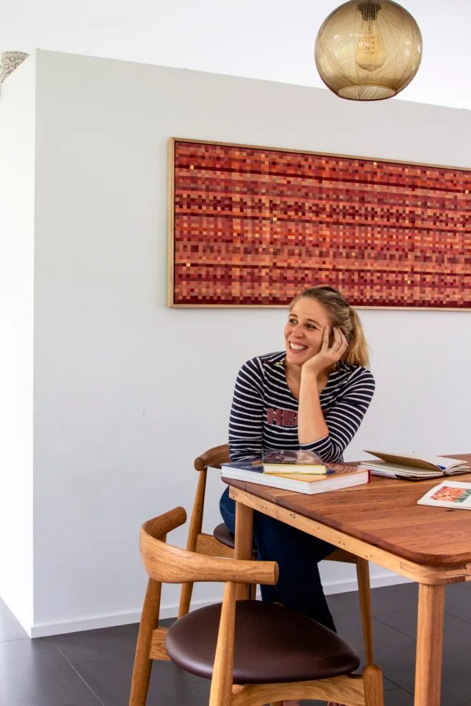 Artist Hannah Cooper sitting a table with a red weave artwork behind her.