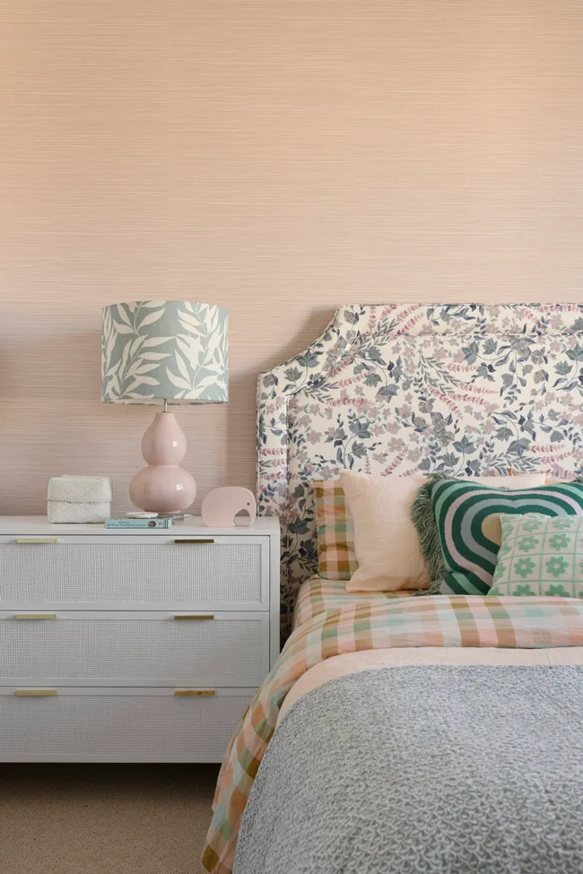 A pastel kids bedroom with patterned linen, white woven drawers and a pink lamp.