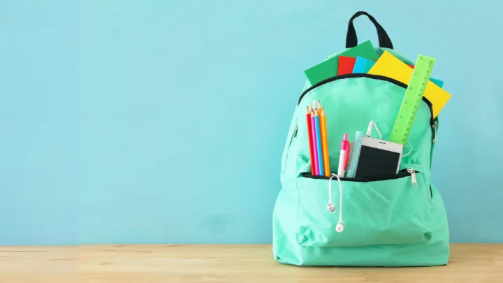 A mint green backpack filled with colorful notebooks, pens, a smartphone, and a ruler against a light blue background.