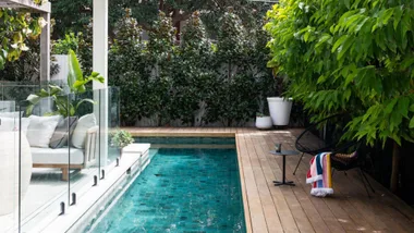 Outdoor pool with wooden deck, glass fence, lush greenery, a lounge chair, and a side table with colorful towels.