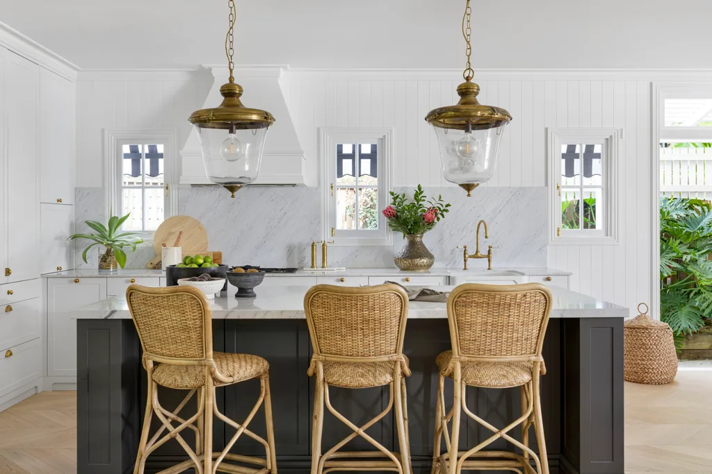 A white and marble French-inspired kitchen with an island, pendant lighting and rattan chairs.