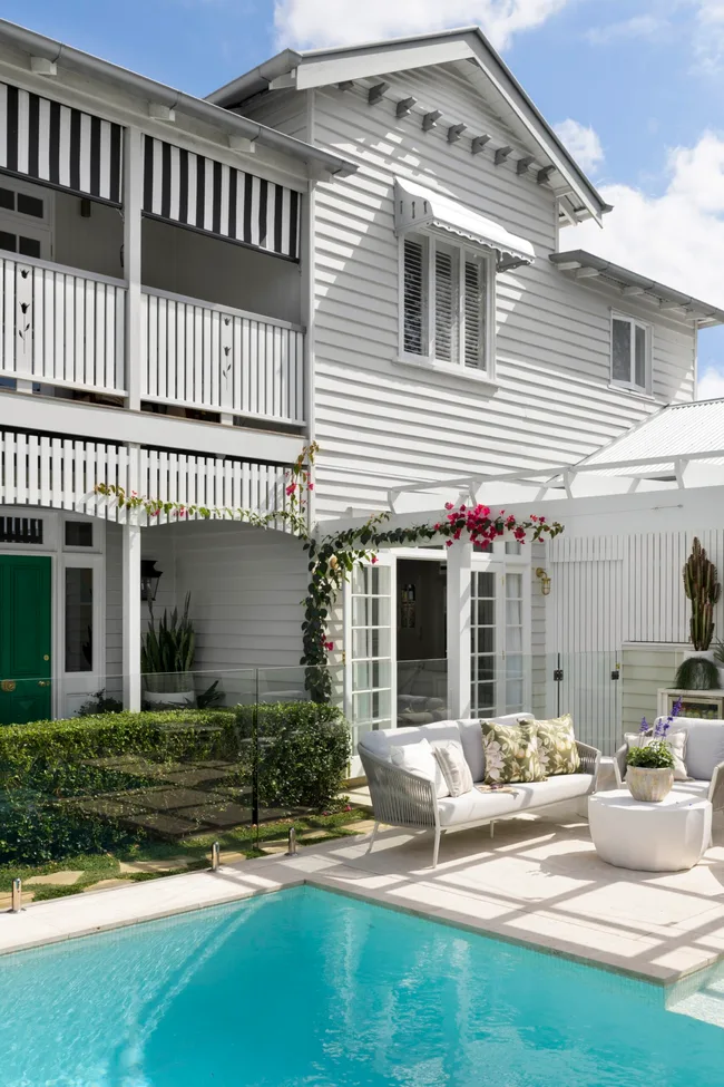 A white two-storey weatherboard cottage beside a pool and lounge chairs.
