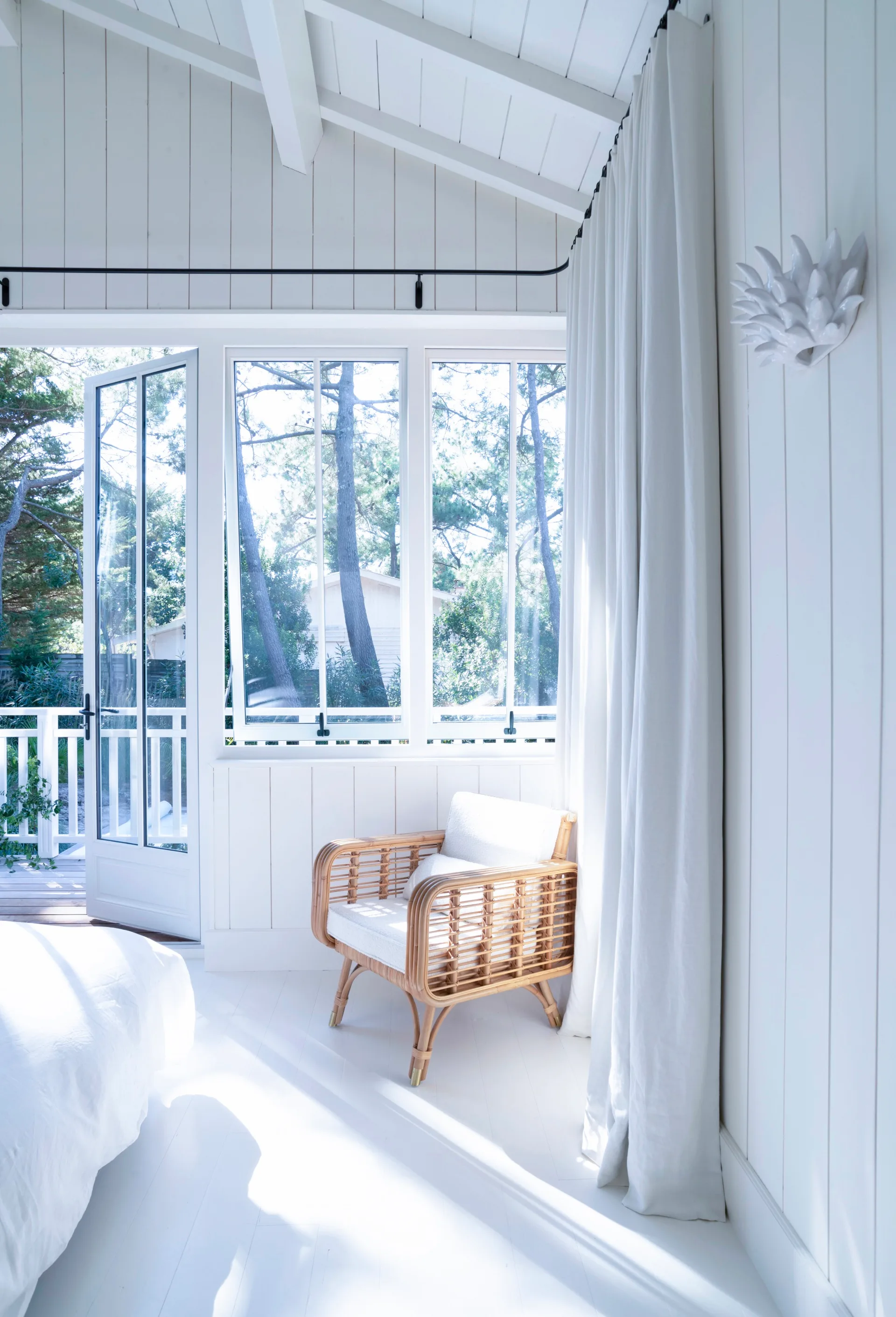 A coastal white bedroom with a wicker chair, wall panels and curtains.