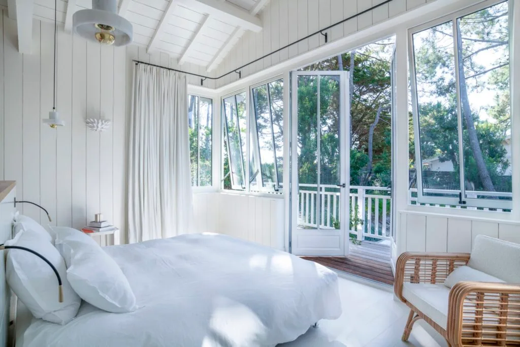 A white bedroom with a bed, wicker chair and balcony deck overlooking a garden.