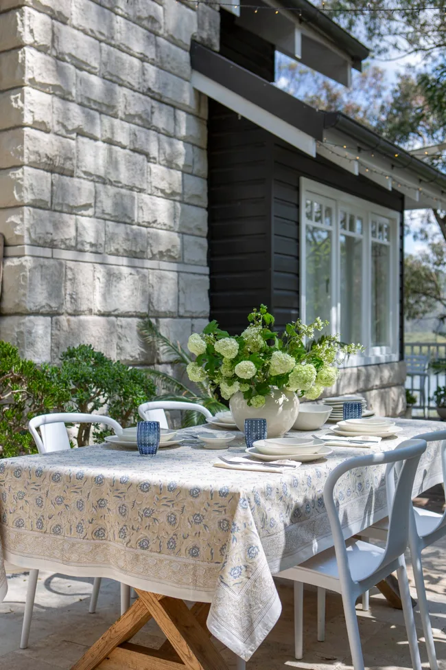 Outdoor table set for dining with floral tablecloth, white chairs, and a large vase of green and white flowers.