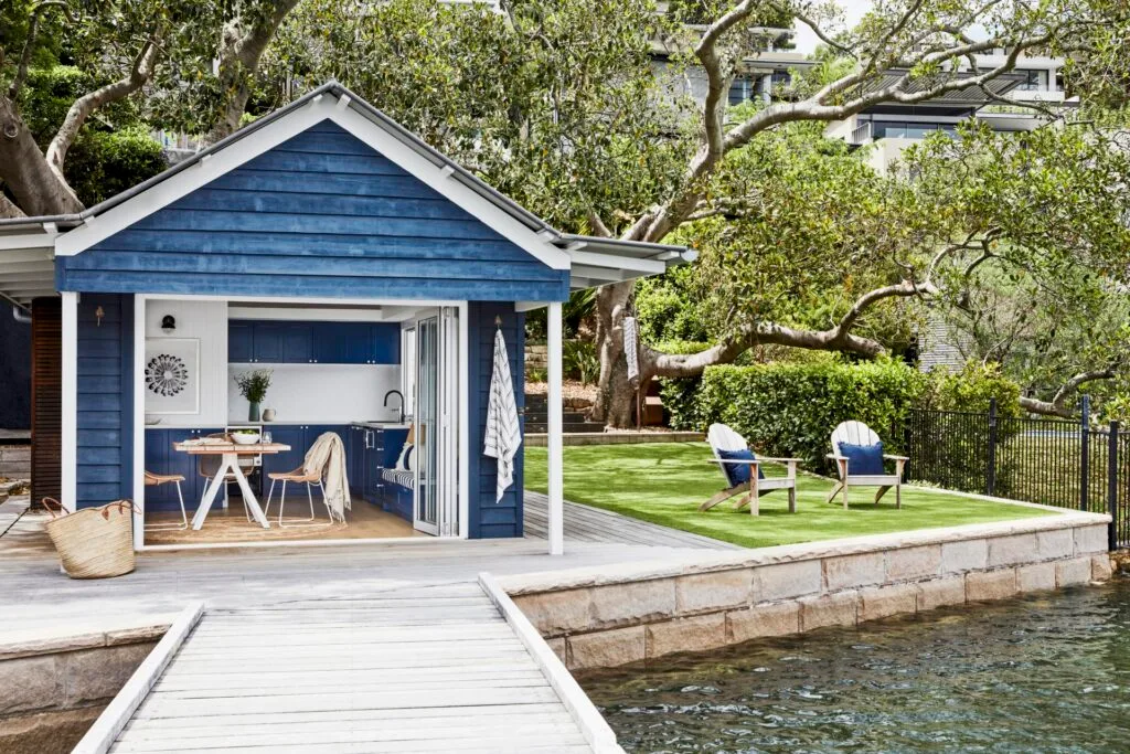 The blue cladded boatshed and two deck chairs on a lawn facing the bay.