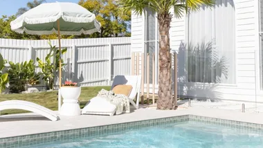 Coastal all white home exterior with sage green umbrella, sun lounger and tiled swimming pool