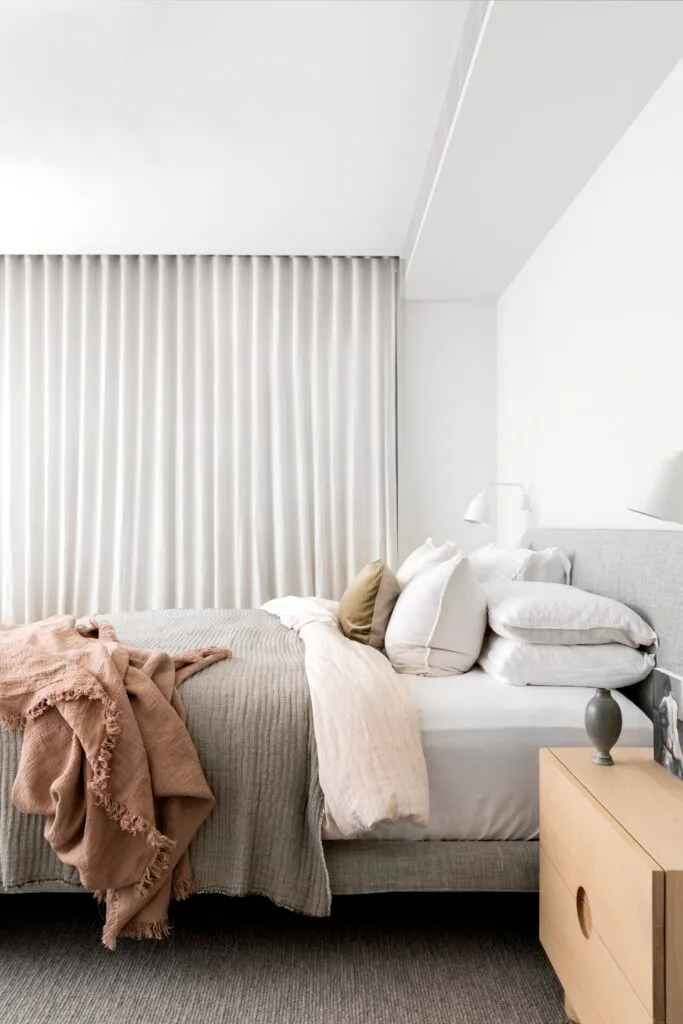 A neutral bedroom with a timber side table, white curtains and grey furnished bed.