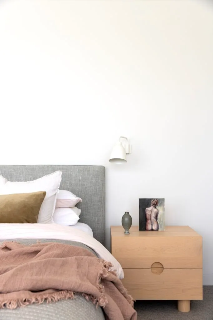 A bed with a grey bedhead, timber table and white sconce.