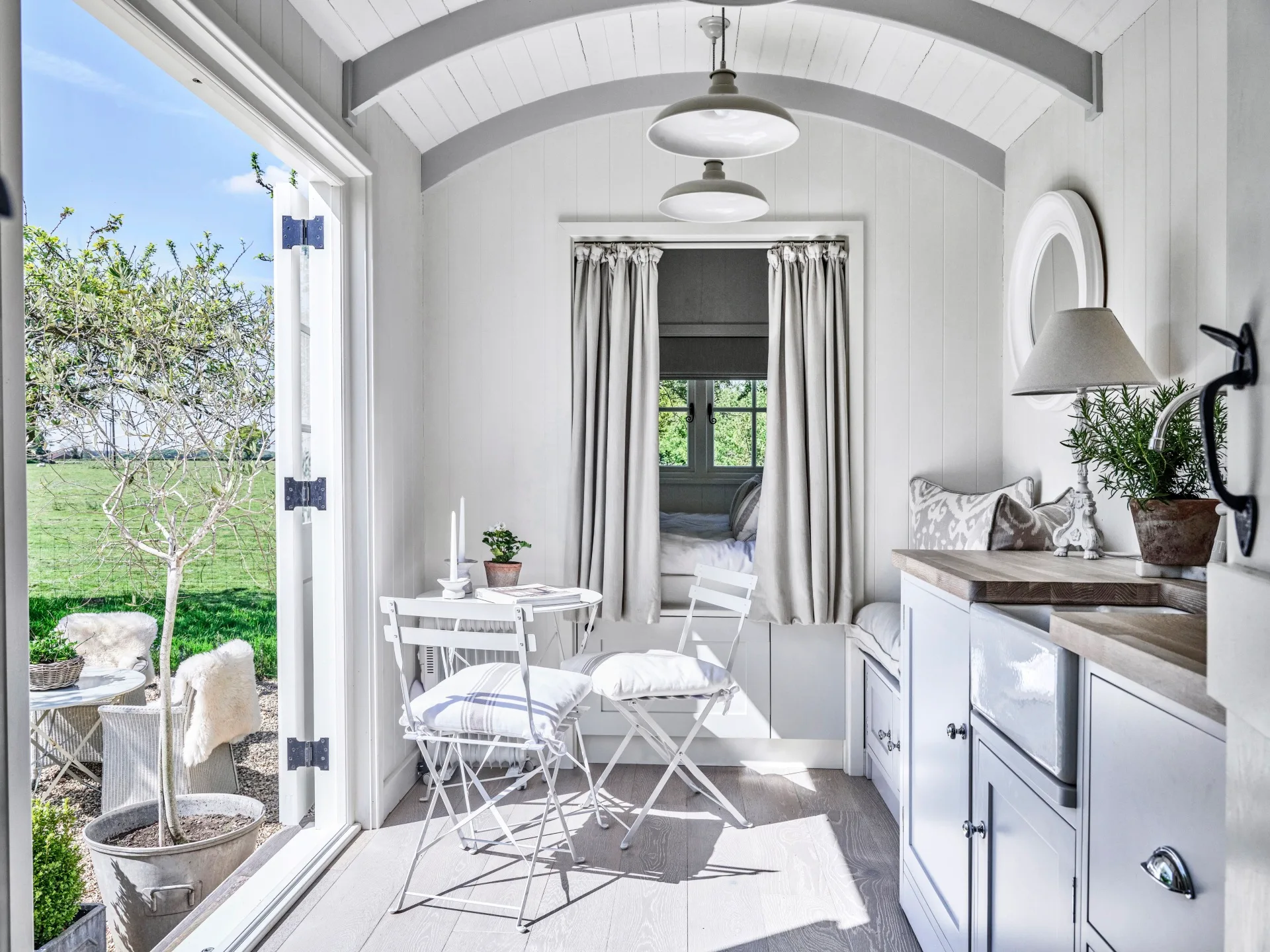 A modern white shepherd's hut with a curtained bedroom and chairs.
