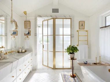 An all-white ensuite with gold glass doors, a freestanding bath and a marble vanity.
