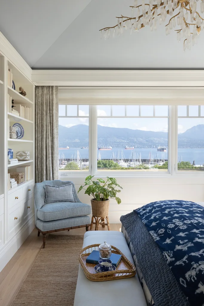 A white, blue and grey main bedroom with windows overlooking a bay.