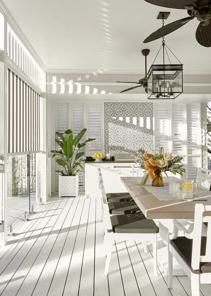 A classic, all-white barbeque deck area with a table, patterned kitchenette tiling and slatted blinds.