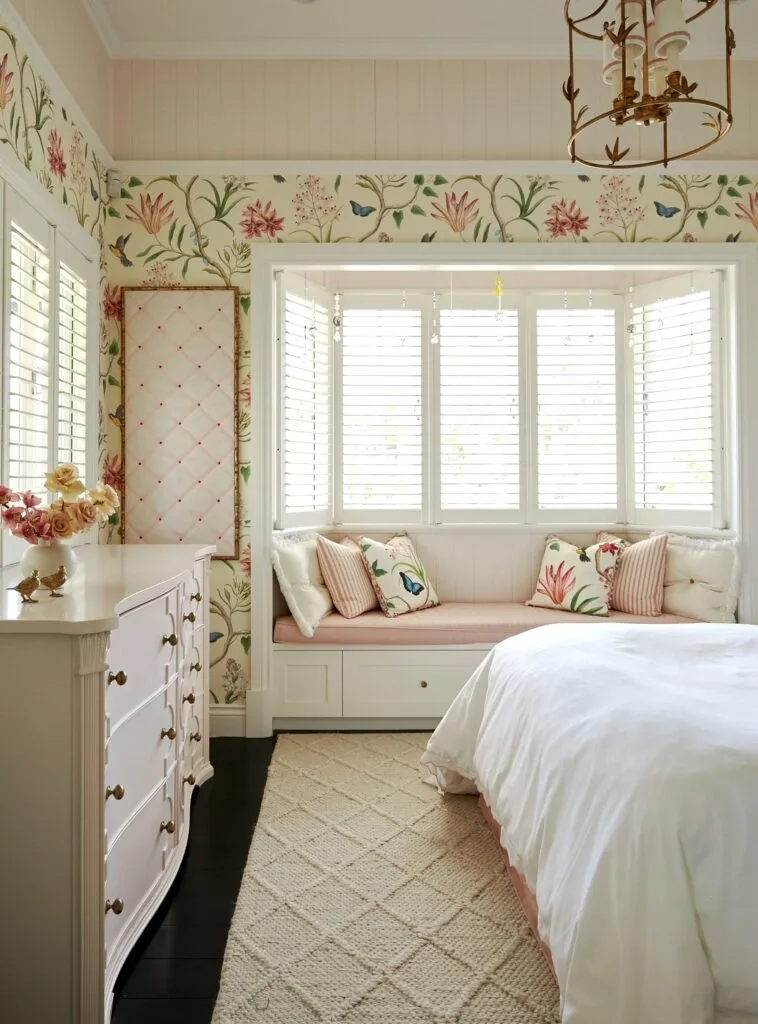 A pink children's bedroom with floral wallpaper, a window seat and vintage style drawers.