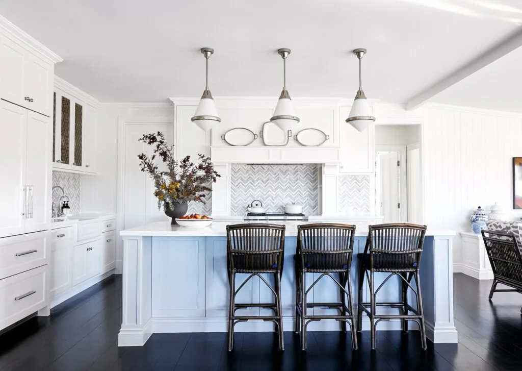 A white classic-style kitchen with shaker cabinets, cane stools, and chevron tiles.