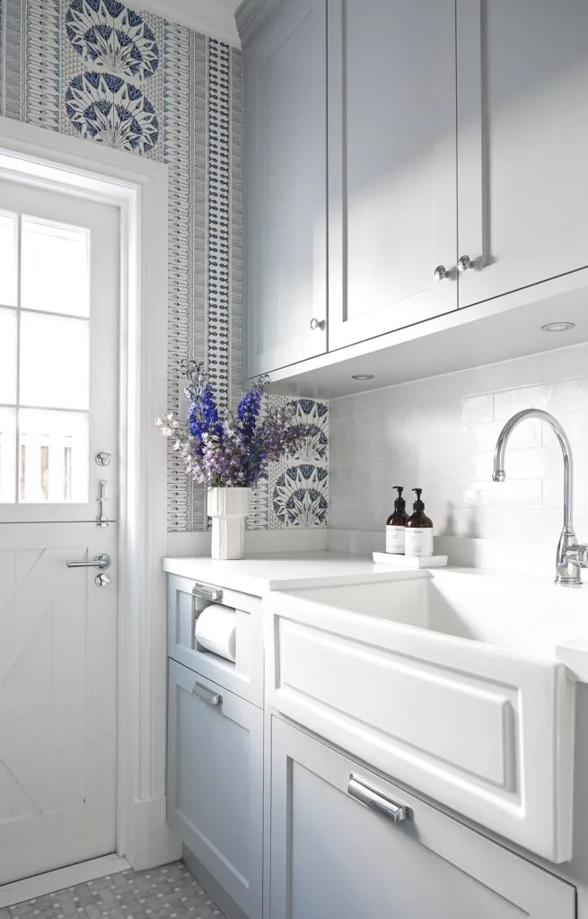 A laundry room with blue wallpaper, shaker cabinets and farm-style sink.