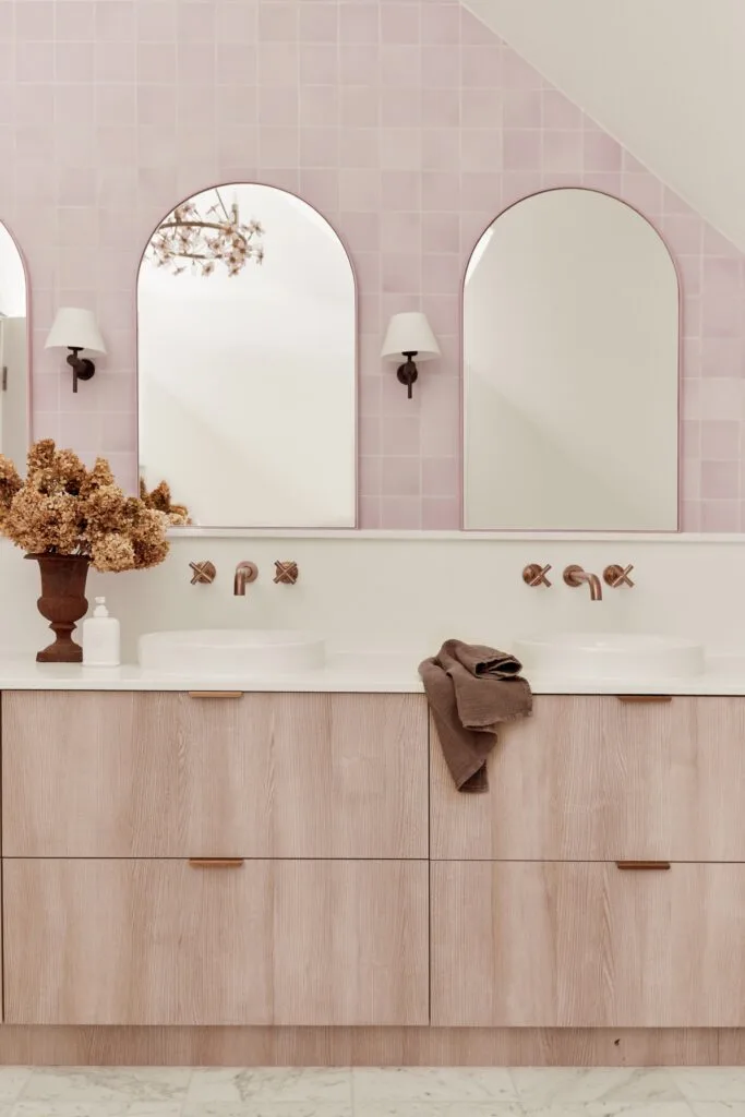 A pastel pink bathroom with a timber vanity, arched mirrors and square pink wall tiles.