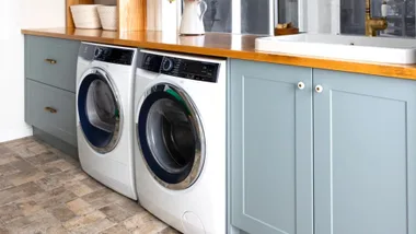 Laundry room with a washing machine and dryer under a wooden countertop, adjacent to a sink and light blue cabinets.