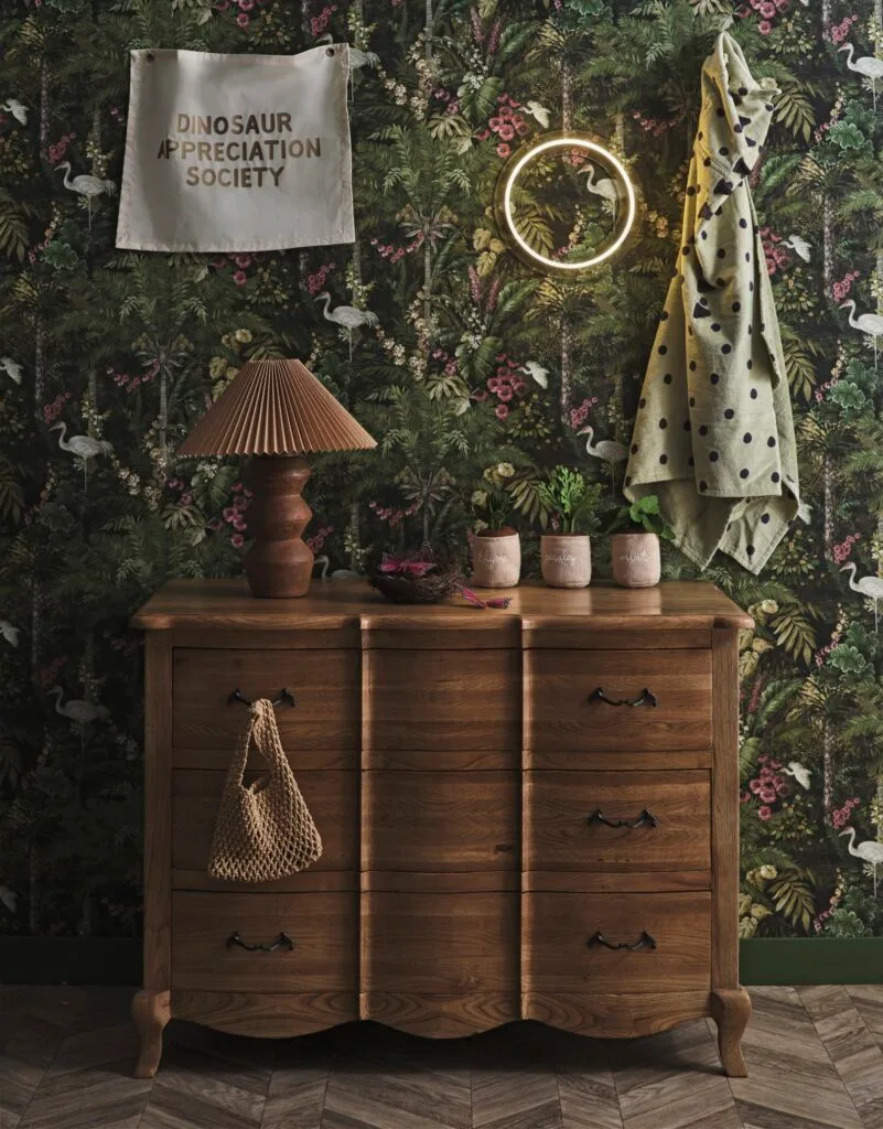 A timber set of drawers in front of green forest wallpaper.