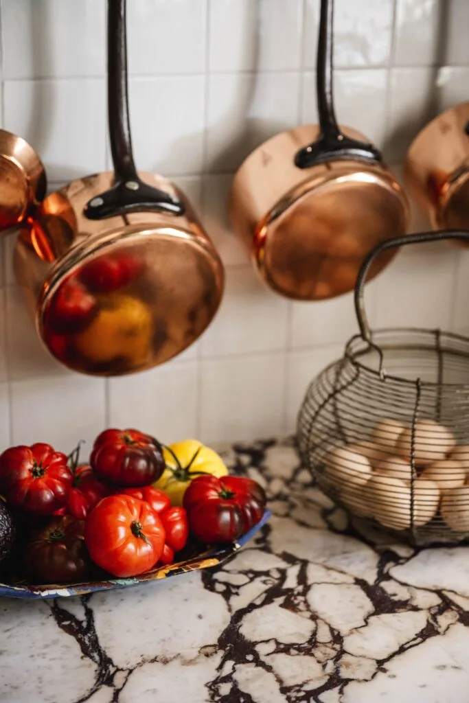 Brass pots hanging above a bowl of tomatoes on a marble bench.