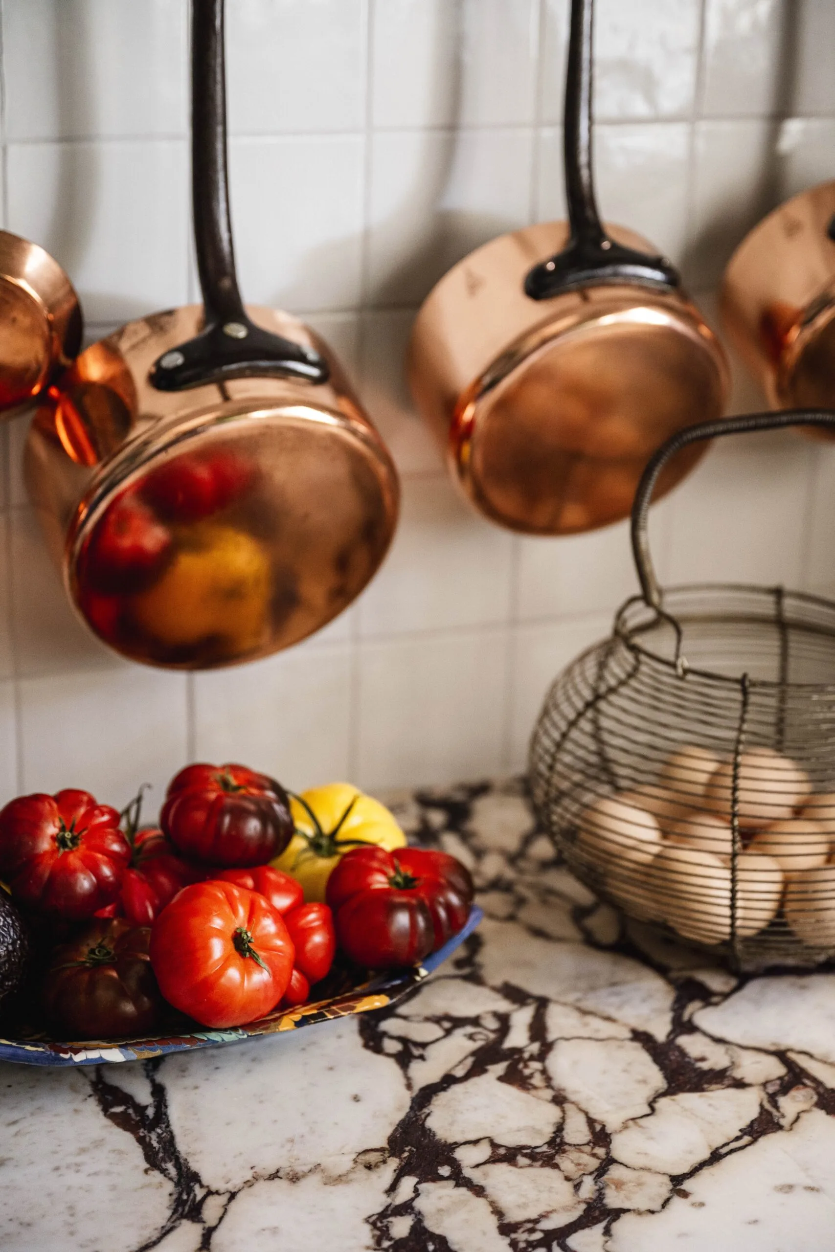 Brass pots hanging above a bowl of tomatoes on a marble bench.