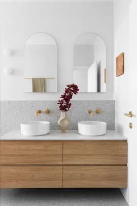 A white and grey ensuite with a timber vanity, gold tapware and arched mirrors.
