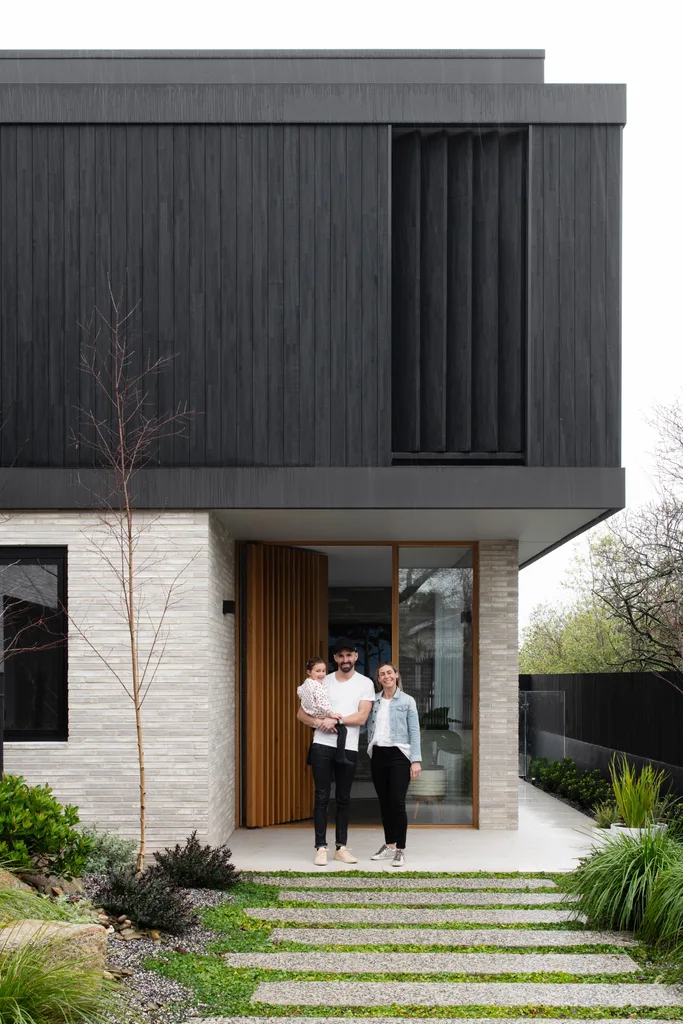 Owner Ed, Jess and Hazel standing at the home's entrance made of stone and black timber cladding.