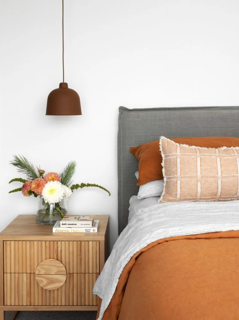 A main bedroom with a grey bed furnished with ochre linen beside a fluted timber table.