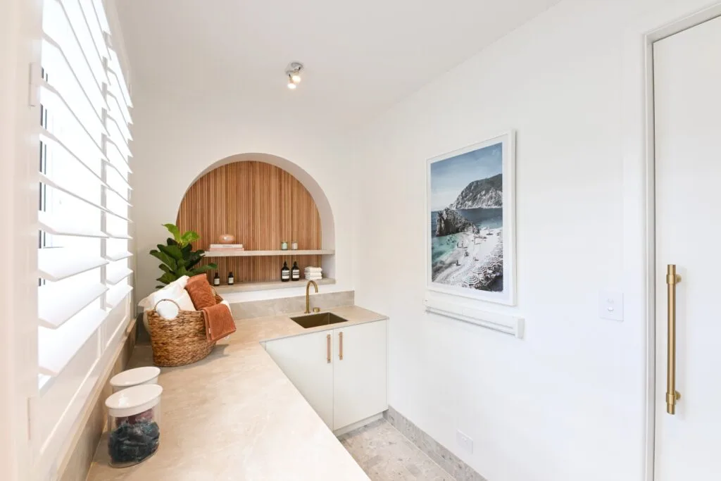 Rhys and Liam's Dream Home laundry with natural coloured benchtop, white cabinetry and gold handles and arch featured in the wall with timber back.