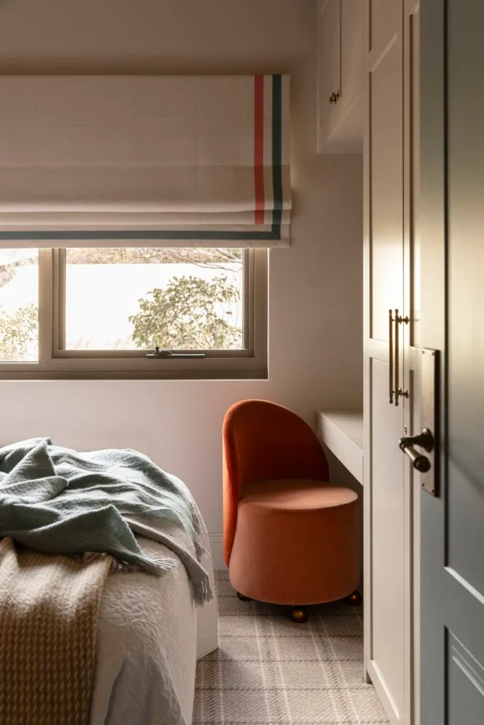 A curved orange chair below a window with white striped blinds.