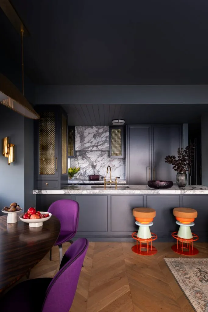 A dark teal kitchen beside a dining nook with purple chairs.