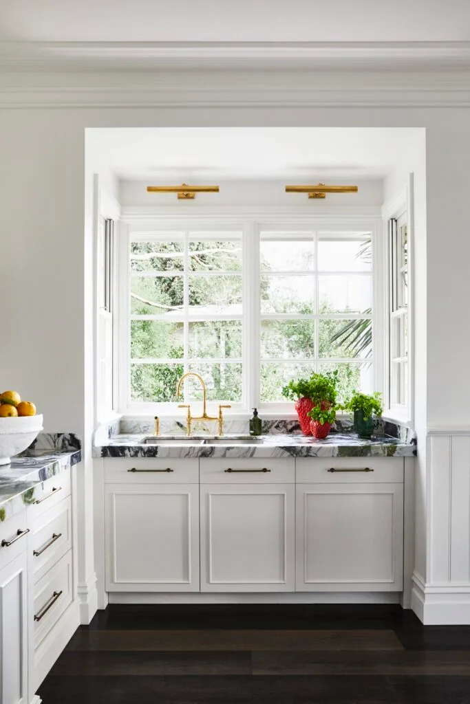 A marble-topped sink with gold tapware beneath double windows.