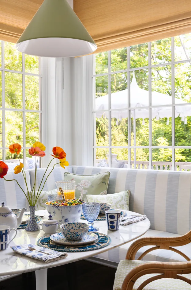 The banquette seating in this breakfast nook makes use of a fabric with blue and white stripes. Red and orange flowers are displayed in a vase atop the oval-shaped table.