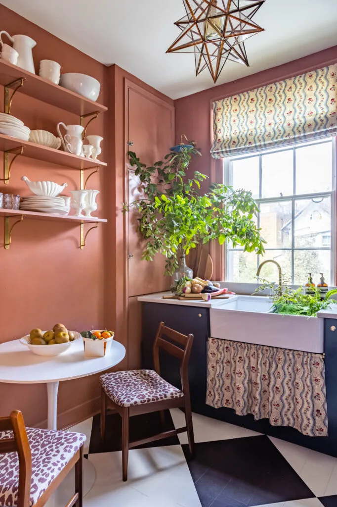 The walls of this kitchen are painted in a bright terracotta shade. Checkerboard flooring provides a pop of pattern underneath a small, circular white table with two dining chairs beside.