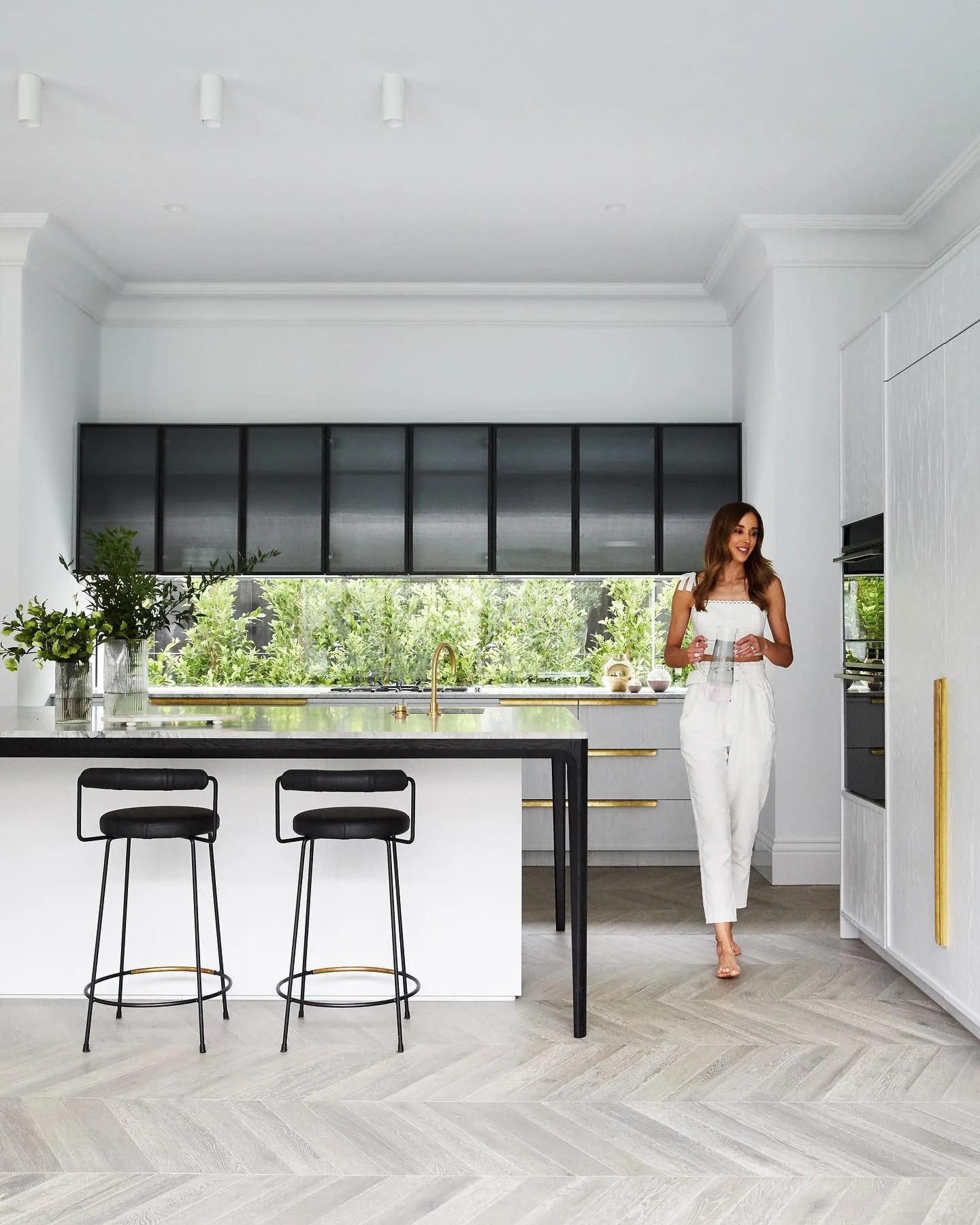 Bec Judd in her kitchen, with brass hardware, black bar stools and cabinetry finished in Judd Soft Grey from Haymes Paint.