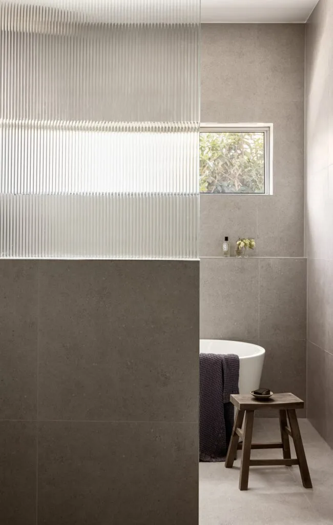 A modern grey bathroom with a fluted glass shower wall and freestanding tub.