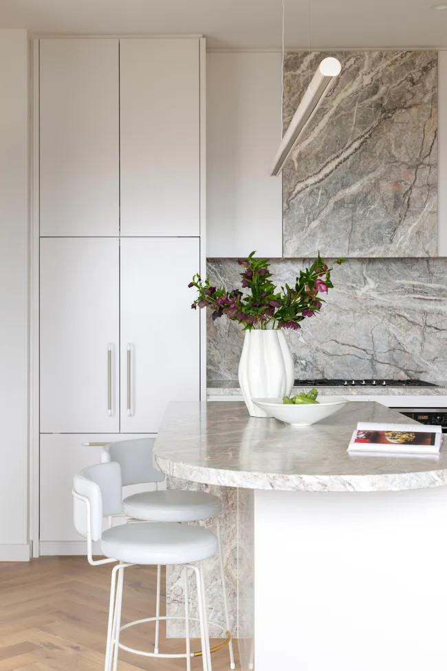 A modern white and marble kitchen with an island and white stools.