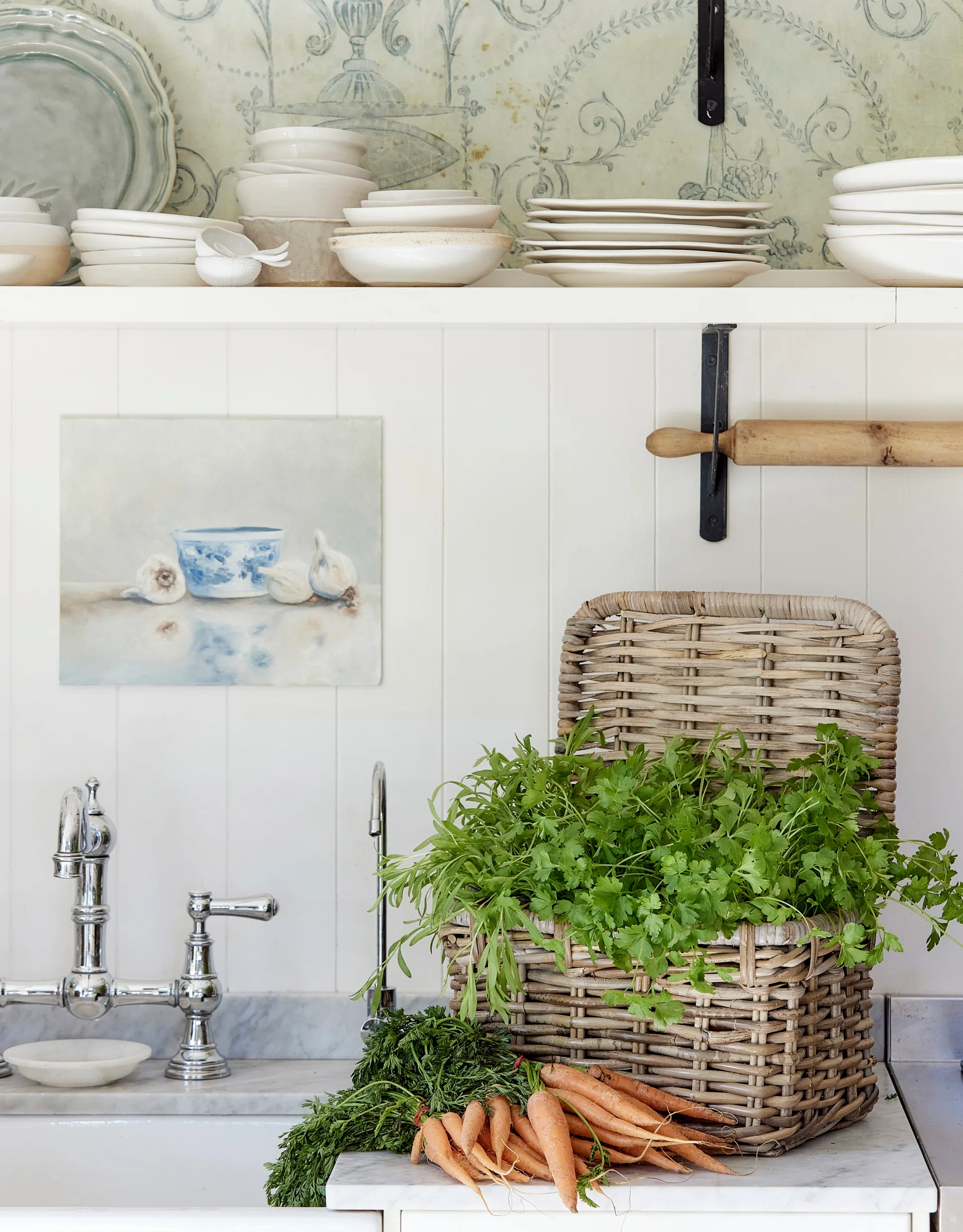 Vertical wooden panels painted in white cover the walls of this kitchen. A woven basket has leafy greens inside with a bundle of carrots beside it. 