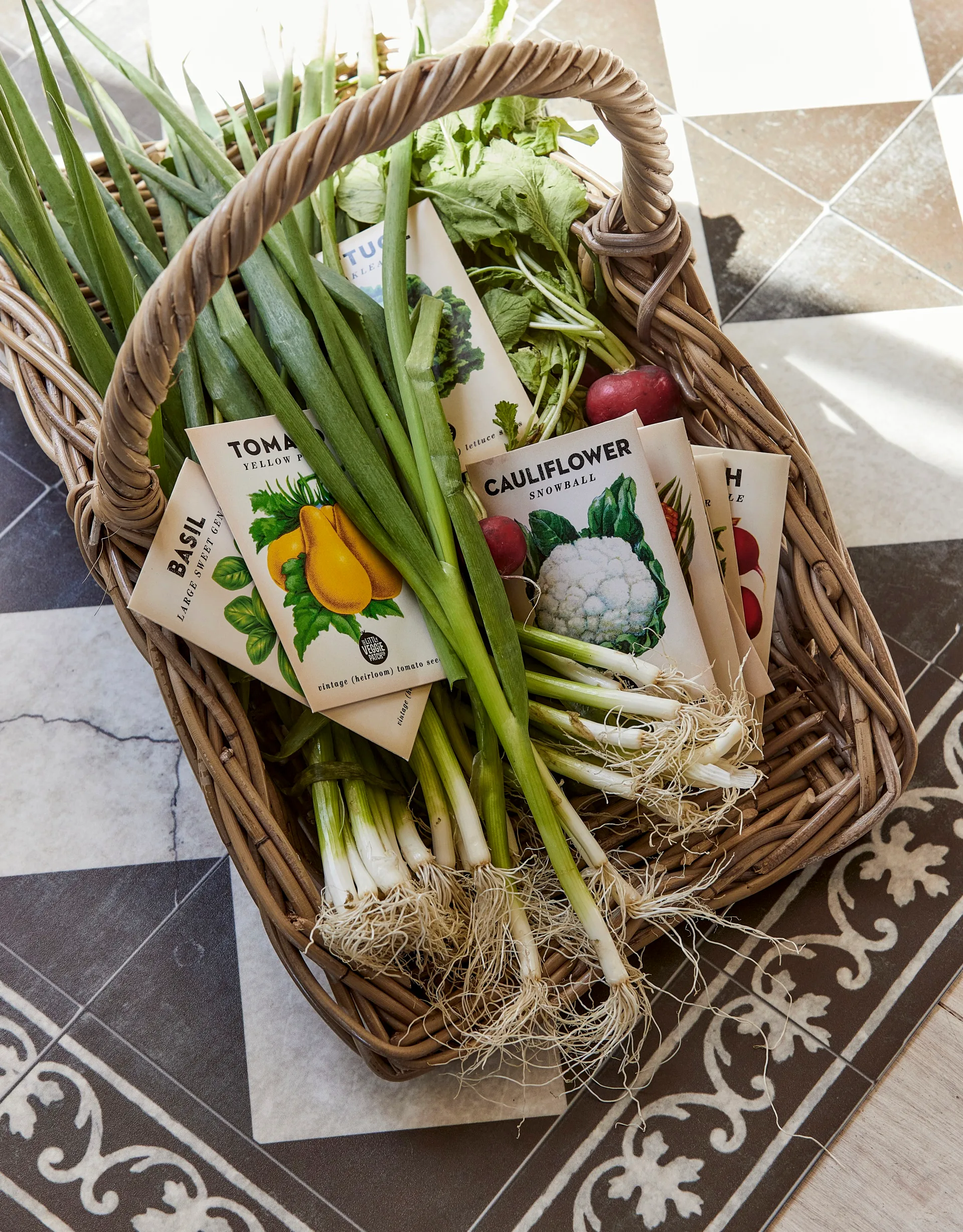 A woven basket is filled with spring onions, radishes and packets of cauliflower, tomato and basil seeds. 