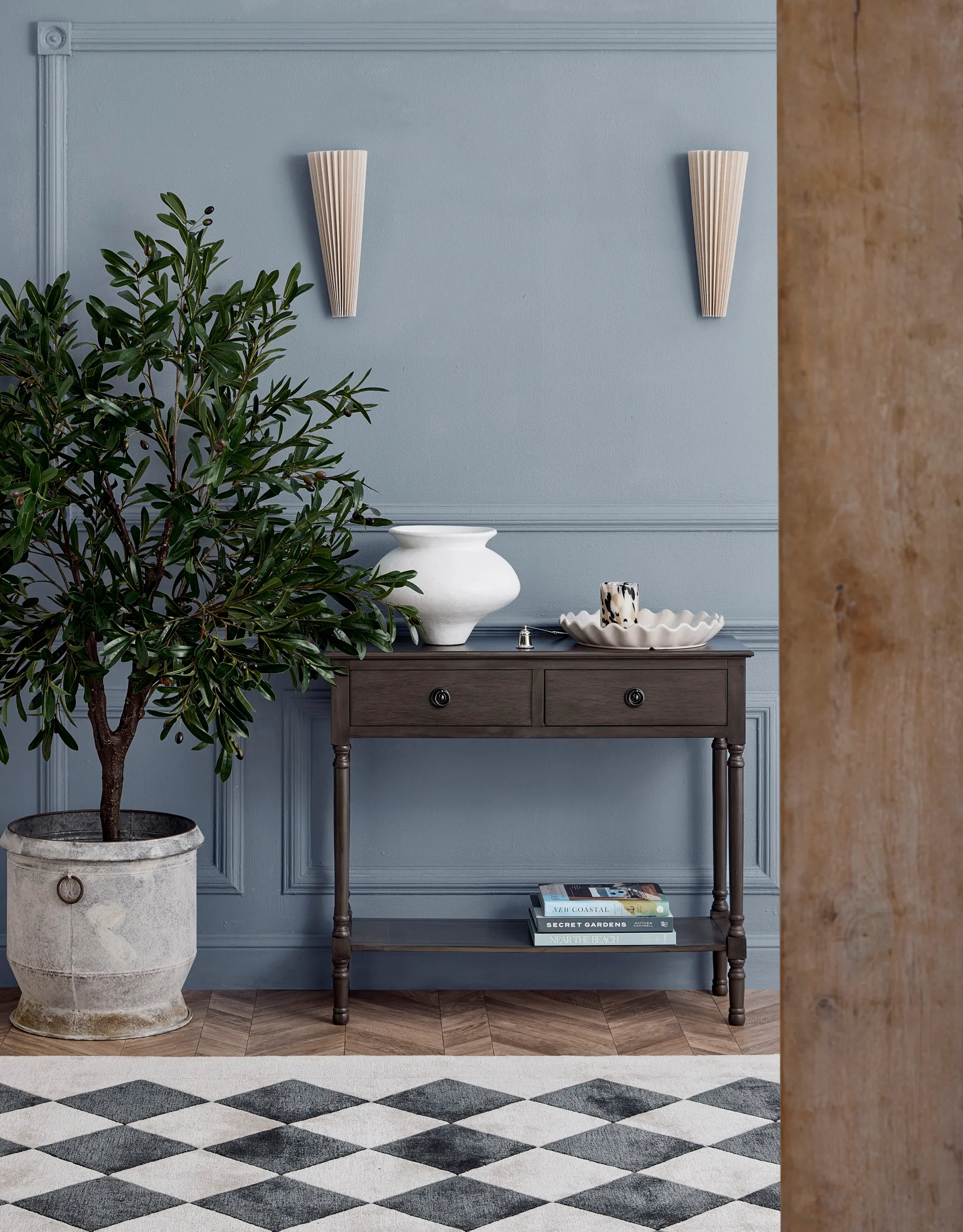 A rug has a diamond pattern in black and white on wooden flooring, against a blue wall with an olive tree in a large pot.