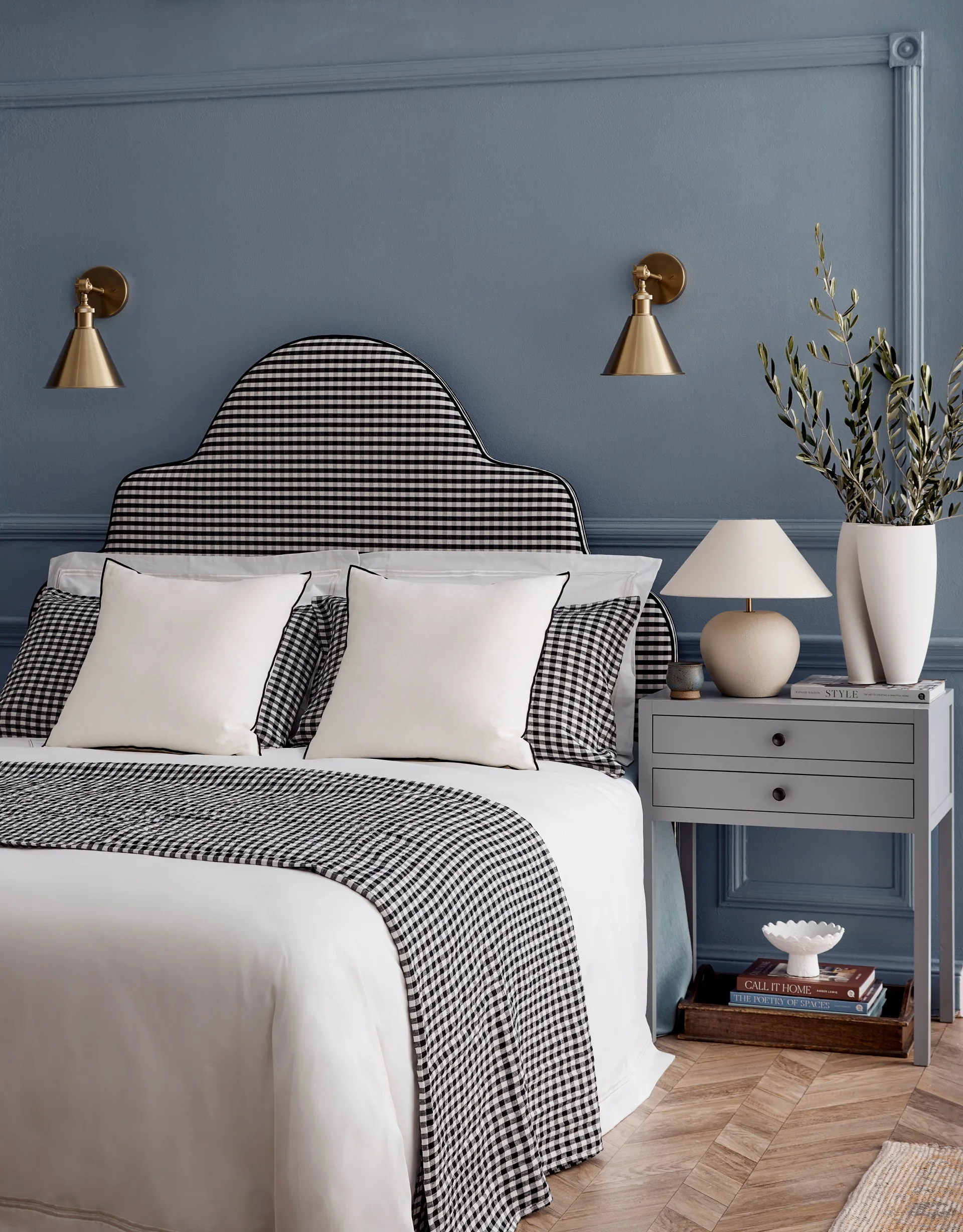 A gingham bedhead, cushions and blanket sit against a blue wall with brass wall lights either side of the bed.