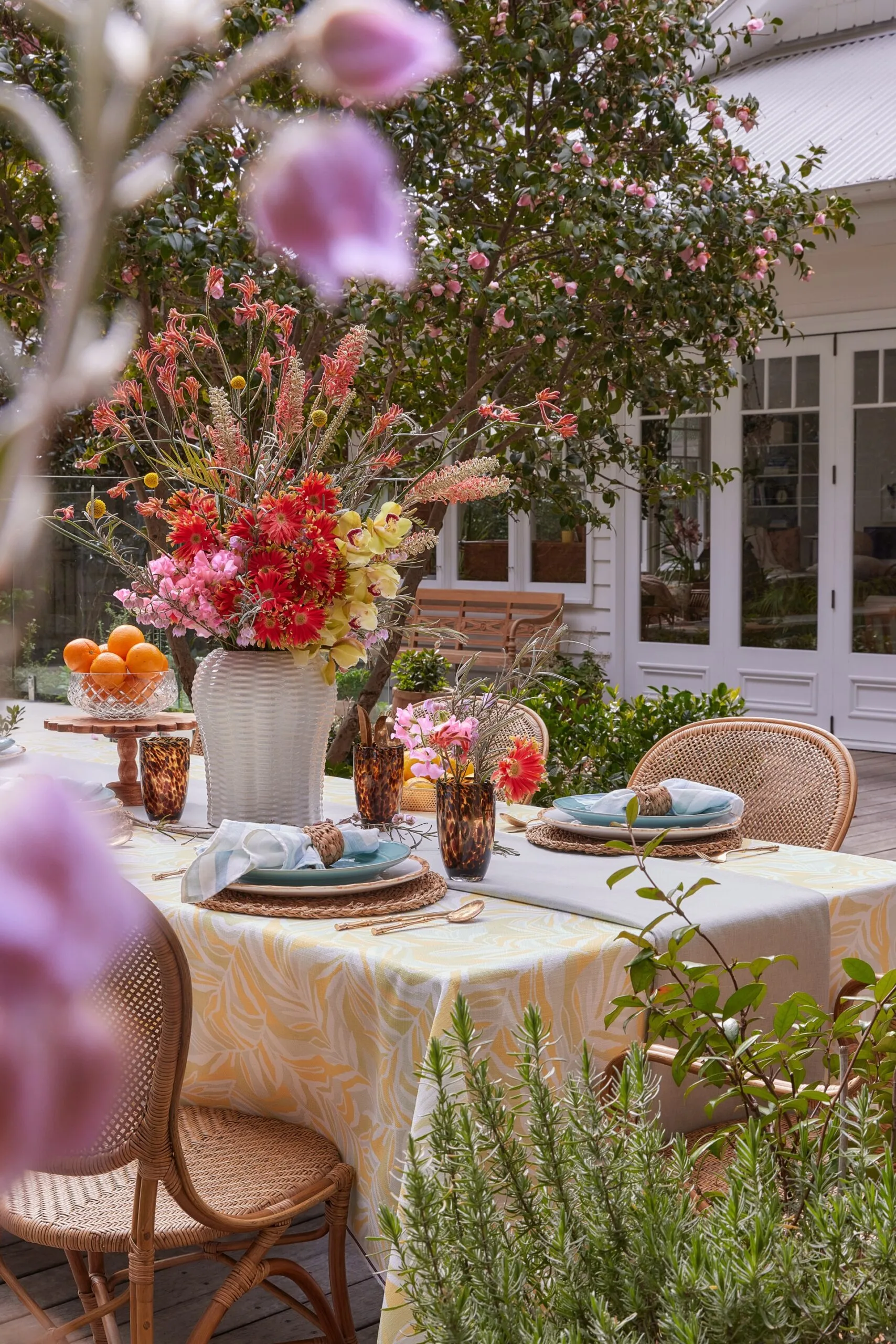 An alfresco dining area with a yellow tablecloth, blue plates, brown glassware and rattan dining chairs.