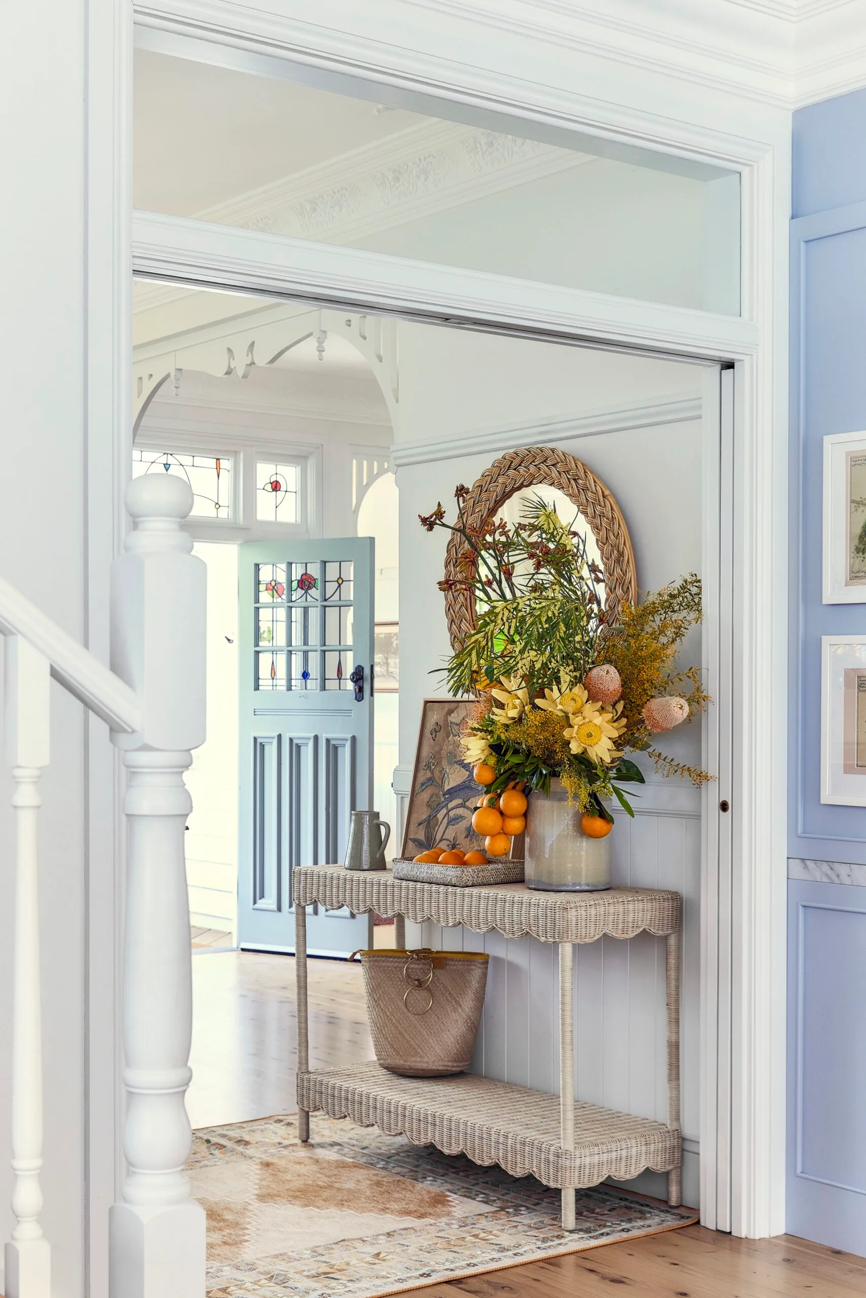 A mirror with a braided frame is positioned above a woven console table, with a blue wall and blue door nearby.