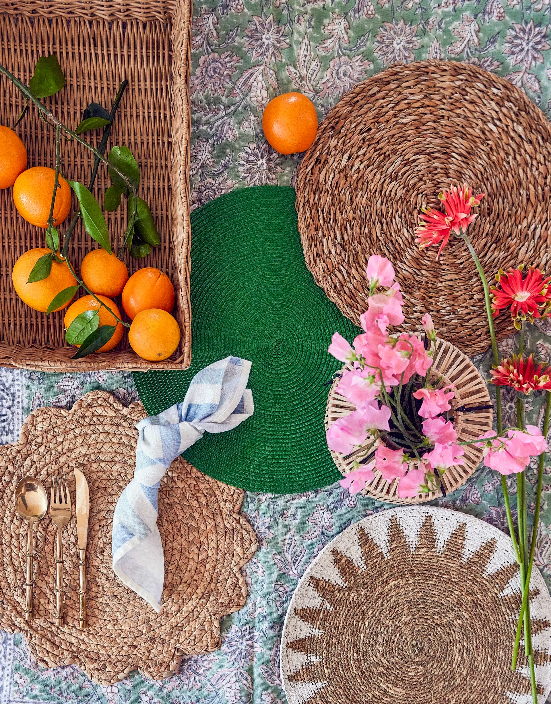 A rattan styling moodboard with woven placemats, a floral tablecloth, oranges and flowers.