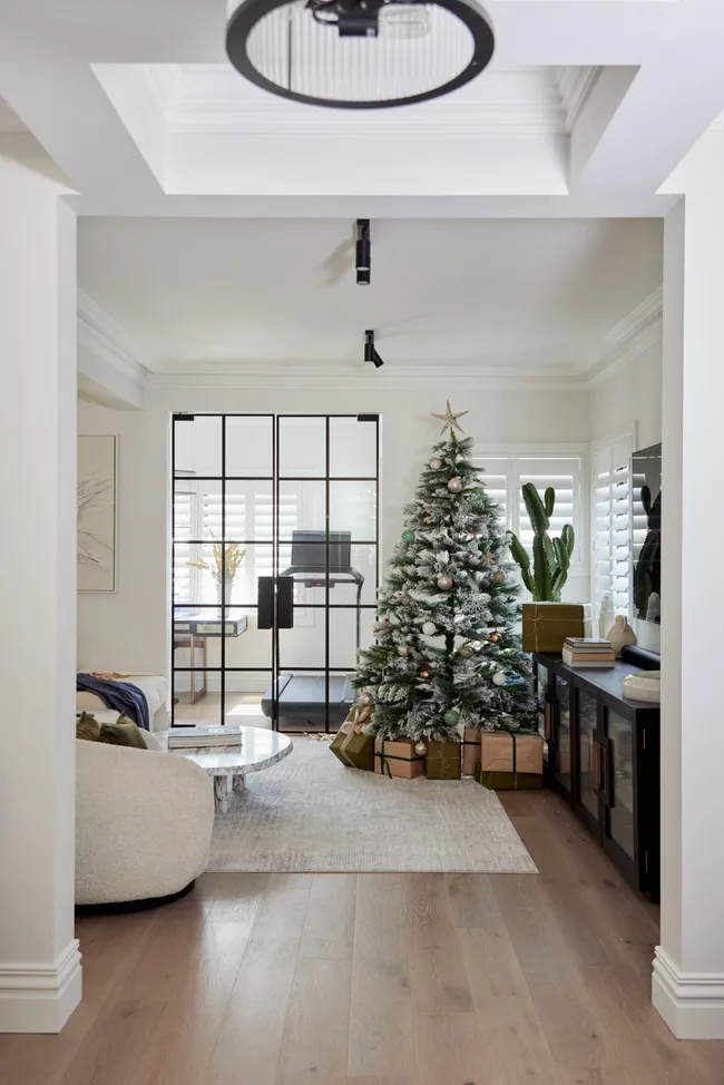 A Christmas tree with faux snow stands proud in a white living room with timber flooring