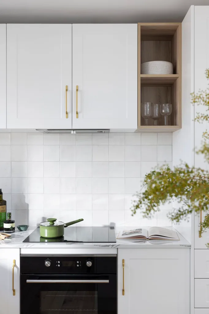 A white tiled splashback wwith white cabinetry and a stovetop with a green pot on it.
